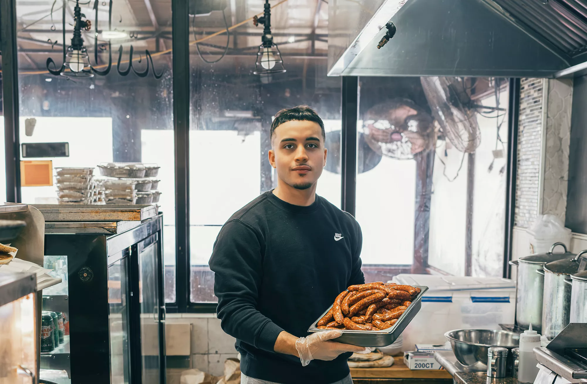 Soufian Mekersi holds a tray of grilled merguez at Merguez and Frites, the café in Queens, New York, that his father opened after emigrating from Algeria. It is known locally for its sausages, french fries and harissa.