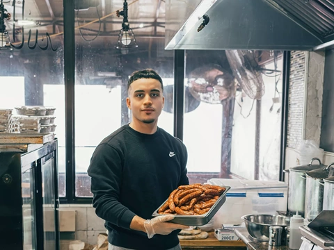 Soufian Mekersi holds a tray of grilled merguez at Merguez and Frites, the café in Queens, New York, that his father opened after emigrating from Algeria. It is known locally for its sausages, french fries and harissa.