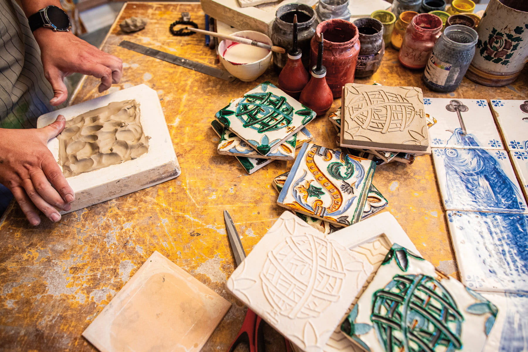 Soft clay is pushed into a square mold to form a tile of three-dimensional design. Other tiles are painted with pigment and metallic paint.