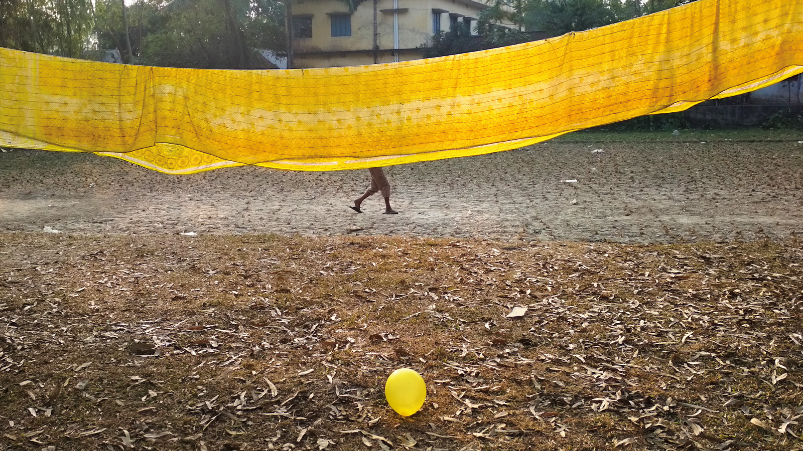 On a winter’s afternoon in 2018 in Bogura, Bangladesh, I went out for a walk with my nephew. While walking, I saw a group of toddlers playing with colorful balloons and across from them boys playing cricket. I was struck to see a bright yellow sari hanging to dry in the sun, which really drew me into to the scene. I noticed that one of the toddlers had a yellow balloon in their hand and I thought about the composition. I positioned myself and waited there for about 10 minutes before the yellow balloon got away from the kids and landed in front of me. I clicked a photo on my cell phone just before one of them ran over to pick it up.
