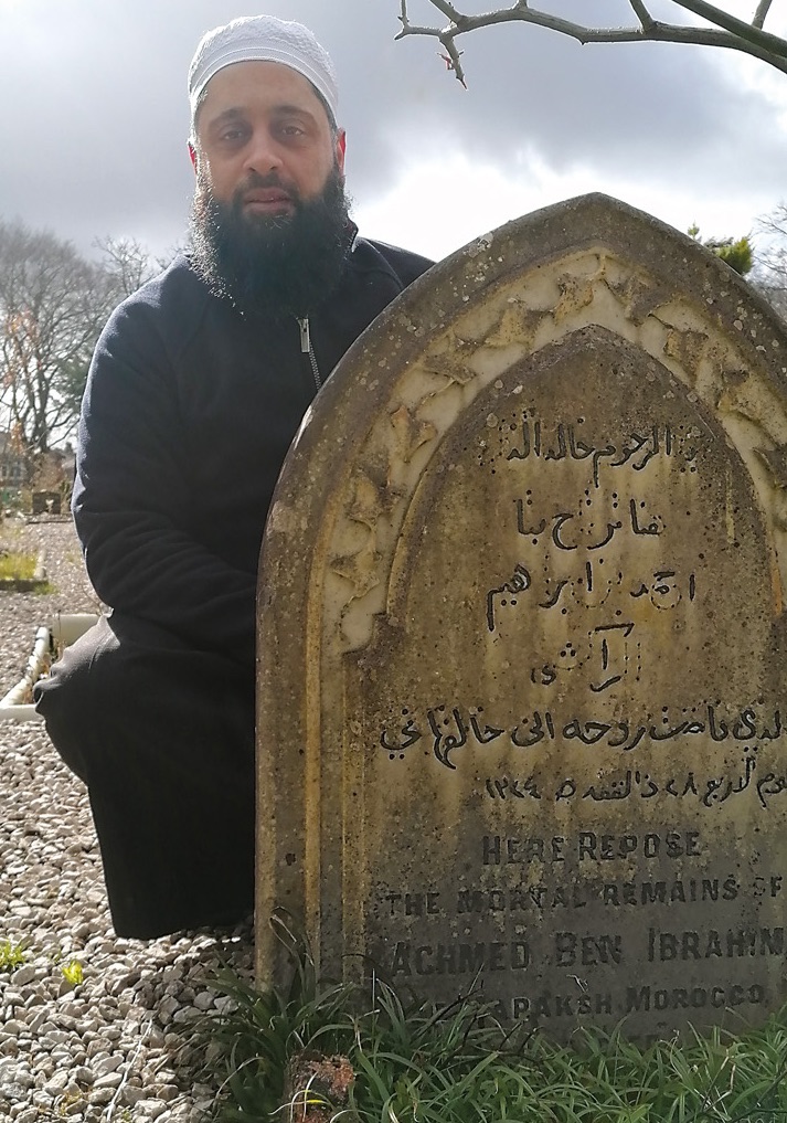 Preston, UK, journalist Ismaeel Nakhuda poses beside the gravestone that gives the acrobat’s full name, Achmed Ben Ibrahim.  