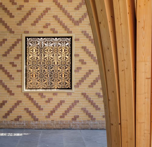 A geometrically patterned screen offers both ventilation and ornamentation in a wall featuring brickwork that incorporates square Kufic calligraphy.