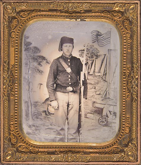 <p>Outfitted with a Zouave fez and equipped with an 1862 Zouave-style sword bayonet, this unidentified Union soldier posed for a portrait against a backdrop depicting a Union military camp</p>
