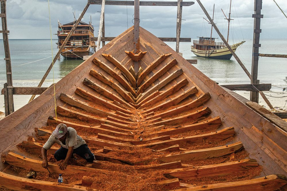 A craftsman scrapes sawdust where ribs meet hull planks on a pinisi boat under construction in Tana Beru